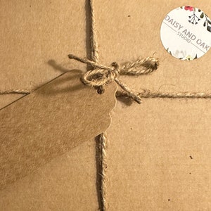 K&ouml;nnte beinhalten: Ein Paket, verpackt in braunem Papier, mit Bindfaden und einer Schleife verschn&uuml;rt. Ein leeres braunes Etikett ist angebracht. Ein runder Aufkleber mit dem Text "DAISY AND OAK STUDIO" und floralen Details ist ebenfalls sichtbar.