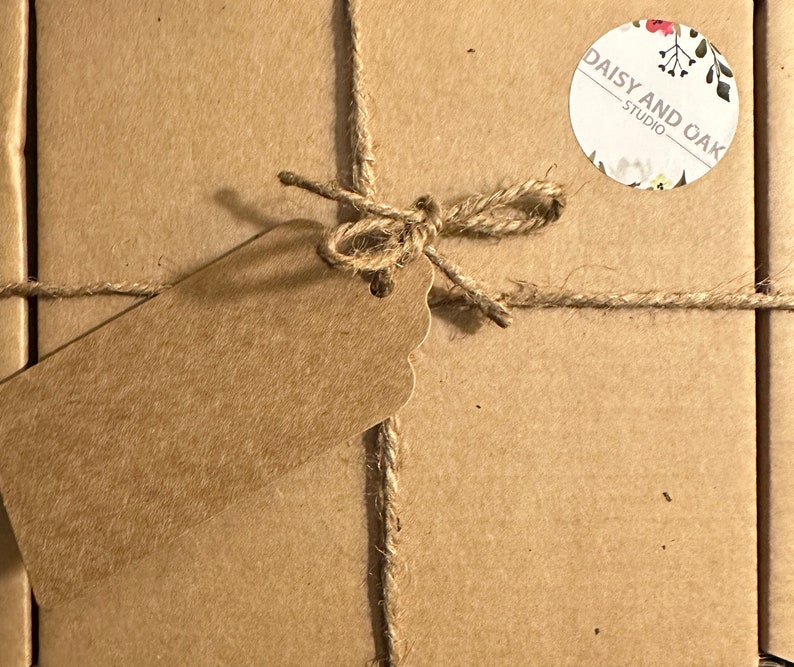 K&ouml;nnte beinhalten: Ein Paket, verpackt in braunem Papier, mit Bindfaden und einer Schleife verschn&uuml;rt. Ein leeres braunes Etikett ist angebracht. Ein runder Aufkleber mit dem Text "DAISY AND OAK STUDIO" und floralen Details ist ebenfalls sichtbar.