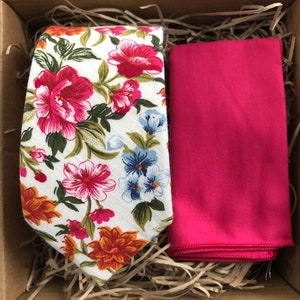 May include: A floral patterned tie and a solid pink pocket square. The tie has a white background with pink, orange, and blue flowers. The pocket square is a solid pink colour.