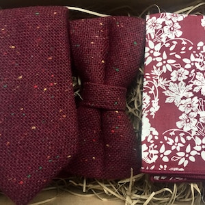 May include: A set of three burgundy accessories for men, including a tie, bow tie, and pocket square. The tie and bow tie are made of a textured fabric with small white and green flecks. The pocket square has a white floral pattern on a burgundy background.