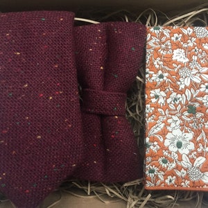 May include: A set of three burgundy and brown patterned accessories for men. The set includes a tie, a bow tie, and a pocket square. The pocket square has a white floral pattern on a brown background.