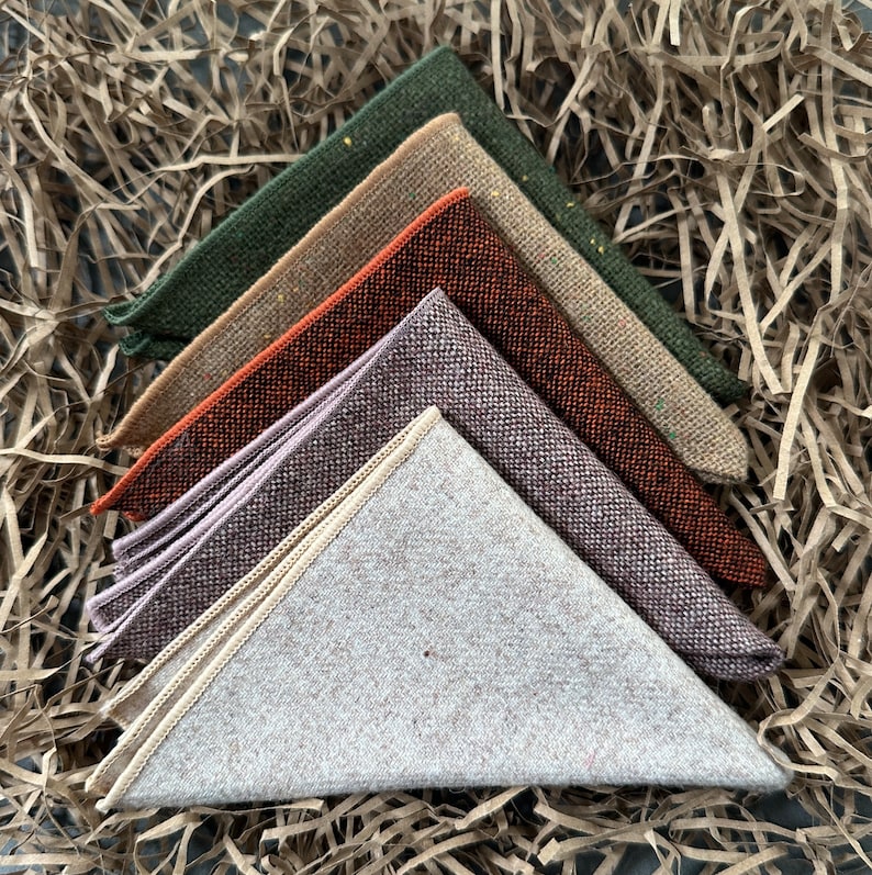 K&ouml;nnte beinhalten: Sechs gefaltete Einsteckt&uuml;cher in verschiedenen Farben, darunter Gr&uuml;n, Beige, Orange, Lila und Grau. Jedes Tuch hat eine strukturierte Webart und einen kontrastierenden Rand. Die Einsteckt&uuml;cher sind f&auml;cherf&ouml;rmig angeordnet.