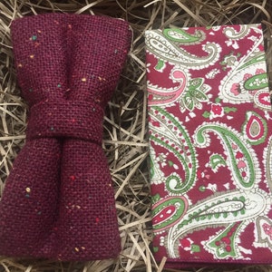 May include: A burgundy bow tie made of burlap with a paisley patterned pocket square. The bow tie has a speckled pattern of red, yellow, and green.
