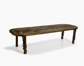 May include: A long, oval-shaped coffee table with a brass top and intricate patterned design. The table has four legs with a unique, wavy design.