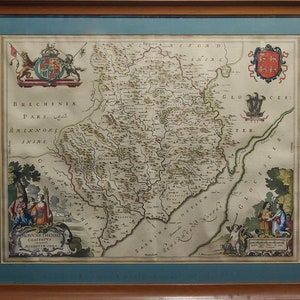 May include: An antique map of Brecknock Shire, Wales, with a detailed depiction of the region's geography and towns. The map is framed in a wooden frame with a blue mat. The map is surrounded by decorative borders and includes a coat of arms in the upper left corner.
