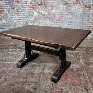 May include: A dark brown wooden table with a thick, rustic top and a unique trestle base. The table has a slightly uneven surface and a distressed finish, giving it a vintage look.