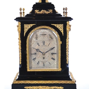May include: A black and gold antique clock with a white face and Roman numeral hour markers. The clock has a decorative design with gold accents and a small, round window on the top.