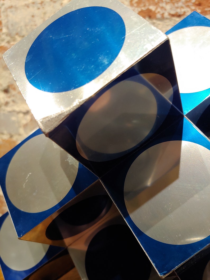 May include: A silver and blue geometric sculpture made of stacked cubes with circular cutouts. Each cube has a blue circle on one side and a silver circle on the other.