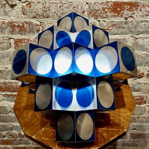 May include: A geometric sculpture made of silver and blue cubes with circular cutouts. The sculpture is arranged in a pyramid shape and is sitting on a wooden surface.