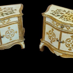 Puede incluir: Par de mesitas de noche de madera pintadas en blanco y dorado con intrincados diseños florales. Cada mesita de noche tiene un cajón y dos puertas.