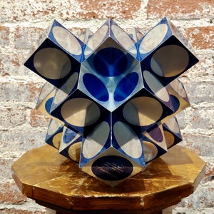 May include: A blue and silver geometric sculpture with a starburst design. The sculpture is made of multiple cubes with rounded edges and is displayed on a wooden pedestal.