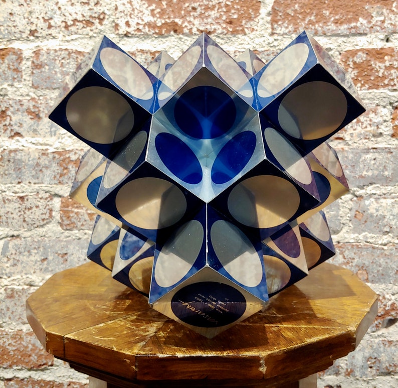 May include: A blue and silver geometric sculpture with a starburst design. The sculpture is made of multiple cubes with rounded edges and is displayed on a wooden pedestal.