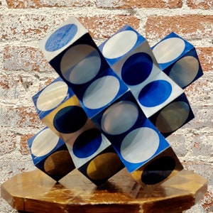 May include: A geometric sculpture made of cubes with alternating blue, white, and gold circles. The sculpture is made of a shiny material and is sitting on a wooden pedestal.
