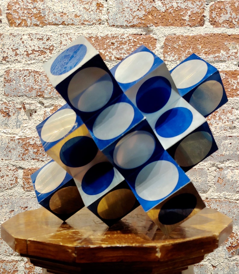 May include: A geometric sculpture made of cubes with alternating blue, white, and gold circles. The sculpture is made of a shiny material and is sitting on a wooden pedestal.