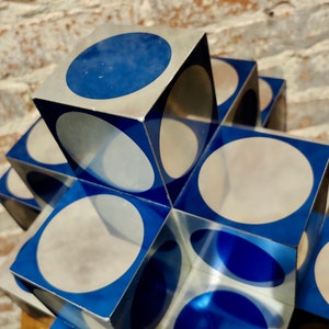 May include: A stack of silver and blue cubes with circular cutouts. The cubes are arranged in a cross shape. The circles are partially visible through the cubes.