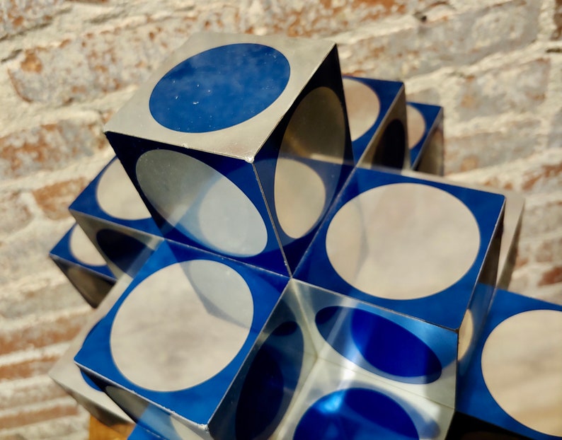 May include: A stack of silver and blue cubes with circular cutouts. The cubes are arranged in a cross shape. The circles are partially visible through the cubes.