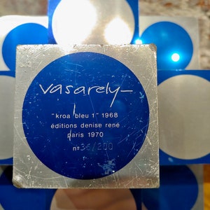 May include: A silver and blue square with a circle in the center. The circle has the text "Vasarely- "kroa bleu 1" 1968 &eacute;ditions denise ren&eacute; paris 1970 №36/200" in black text.
