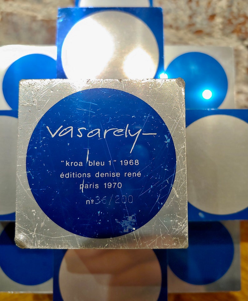 May include: A silver and blue square with a circle in the center. The circle has the text "Vasarely- "kroa bleu 1" 1968 &eacute;ditions denise ren&eacute; paris 1970 №36/200" in black text.