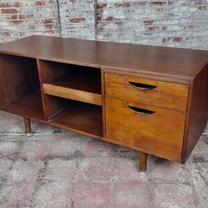 May include: A mid-century modern walnut wood media console with two drawers and open shelving. The console has a dark wood finish and sits on tapered legs.