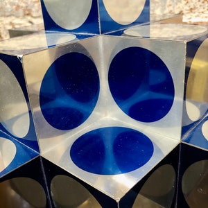 May include: A cube-shaped sculpture made of clear acrylic with blue and silver circles on each side.