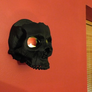 May include: A black skull-shaped candle holder with a mirrored eye socket. The candle holder is mounted on a red wall.