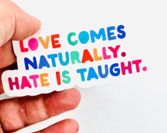 Puede incluir: Una pegatina colorida con la frase "LOVE COMES NATURALLY. HATE IS TAUGHT." en una fuente juguetona. El texto está en varios colores, incluyendo rosa, naranja, azul y verde. La pegatina se sostiene sobre un fondo blanco.