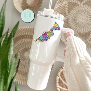 May include: White stainless steel tumbler with a colourful trumpet sticker. The tumbler has a lid and a handle.
