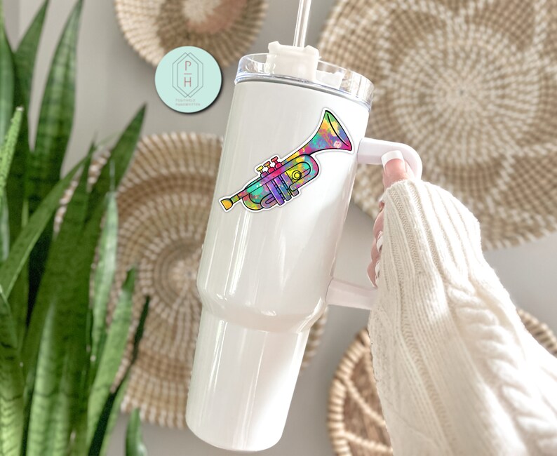 May include: White stainless steel tumbler with a colourful trumpet sticker. The tumbler has a lid and a handle.