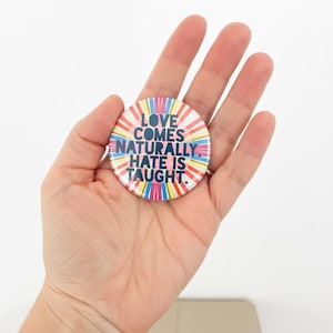 May include: A circular badge with a vibrant, multi-coloured radial pattern. The badge displays the message "LOVE COMES NATURALLY. HATE IS TAUGHT." in a clear, sans-serif font, promoting a message of positivity.