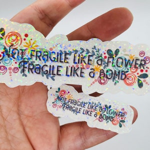May include: Two iridescent glitter stickers with the text "Not Fragile Like a Flower. Fragile Like a Bomb." The stickers have a floral design with colourful swirls and dots.