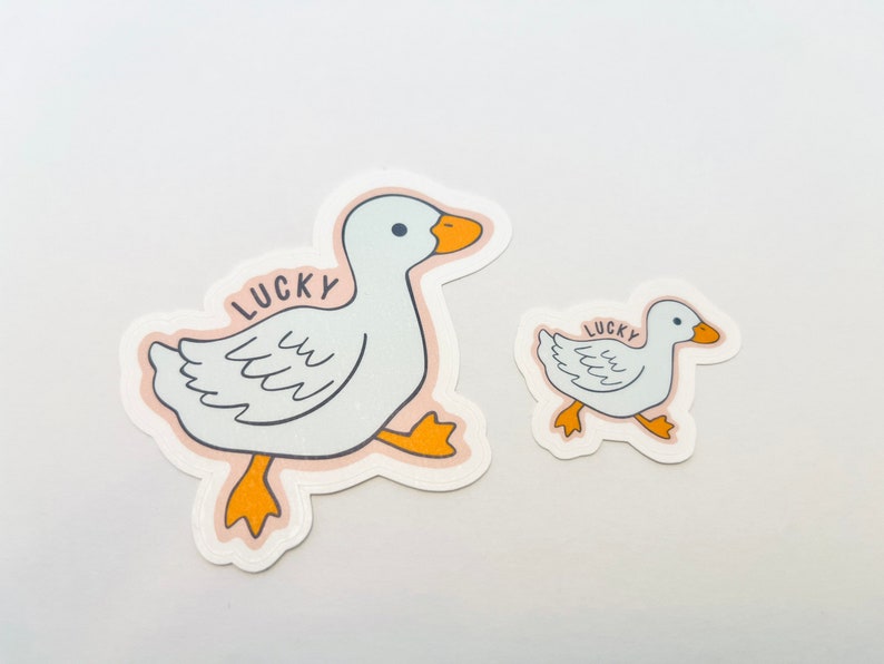 May include: Two white duck stickers with the word "LUCKY" printed on them. The larger duck is walking to the right and the smaller duck is walking to the left.