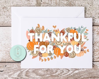 May include: A white greeting card featuring the text THANKFUL FOR YOU in bold white letters. The design includes orange pumpkins, brown and tan circular floral patterns, and muted green leaves arranged in a cluster behind the lettering.
