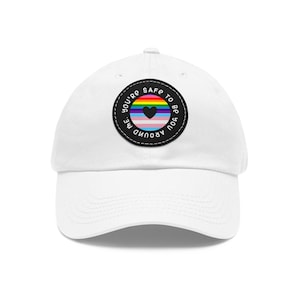 Op de afbeelding: Witte baseballpet met een zwart-witte patch waarop staat "You're safe to be around me" met een regenboog hart en een transgender pride-vlag erin.