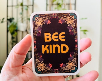 May include: A black and gold sticker with a floral border and the text "Bee Kind" in yellow. The sticker features four stylized bees with brown bodies and yellow wings.