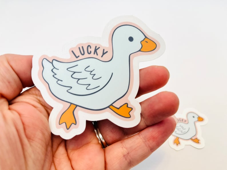 May include: A white duck sticker with orange feet and beak. The sticker has a pink outline and the word "LUCKY" written on it.