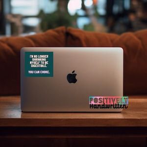 K&ouml;nnte beinhalten: Ein silberner Laptop mit einem schwarzen Apple-Logo. Auf dem Deckel befindet sich ein t&uuml;rkisfarbener Aufkleber mit wei&szlig;em Text, der lautet: "I'M NO LONGER SHRINKING MYSELF TO BE DIGESTIBLE. YOU CAN CHOKE." Darunter befindet sich ein Aufkleber mit der Aufschrift "POSITIVELY Handwritten".