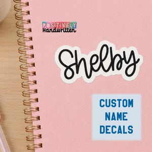 May include: A pink spiral-bound notebook with a gold spiral. The notebook features a sticker with the name "Shelby" in black cursive. A blue sticker reads "Custom Name Decals". The notebook also has a sticker that says "Positively Handwritten".