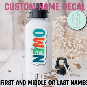 以下が含まれることがあります： 白いウォーターボトルに、虹色の垂直フォントで「Owen」と書かれたカラフルなカスタムネームステッカーが貼られています。ステッカーは、黒い蓋と黒い持ち手付きの白いウォーターボトルに貼られています。ボトルは白い木製の表面の上に置かれています。画像の下部には「FIRST AND MIDDLE OR LAST NAMES」というテキストがあります。