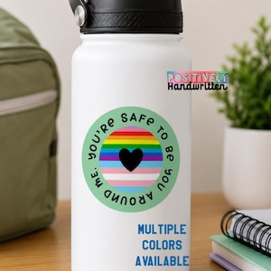 May include: A white water bottle with a black lid and a green sticker. The sticker has a rainbow design, a black heart, and the words "YOU'RE SAFE TO BE YOU AROUND ME." The text "MULTIPLE COLORS AVAILABLE" is also visible.