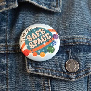 May include: A round button with the text "THIS IS A SAFE SPACE" in a retro space-themed design. The button features planets, stars, and orange and teal accents. It is pinned to a denim jacket.
