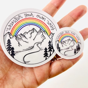 May include: Two round stickers with a mountain range, rainbow, and the text "wander your own way". The larger sticker is white with a black outline and the smaller sticker is white with a black outline.