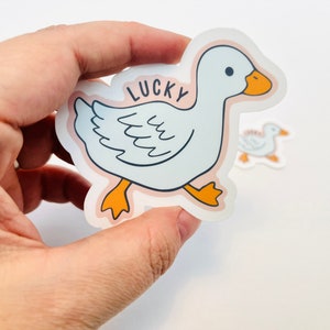 May include: A white duck sticker with orange feet and a pink background. The duck is walking and has the word "LUCKY" written on its back.