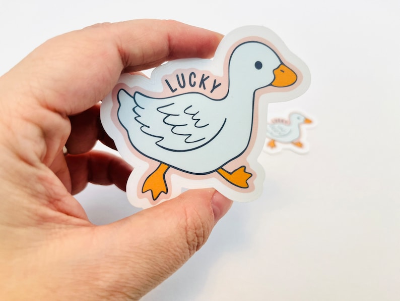 May include: A white duck sticker with orange feet and a pink background. The duck is walking and has the word "LUCKY" written on its back.