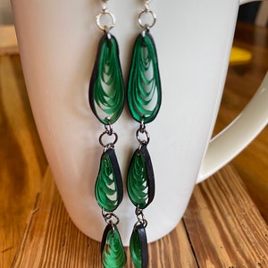 May include: A pair of green and black dangle earrings with three teardrop-shaped paper quilling designs on each earring.