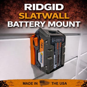 Puede incluir: Una bater&iacute;a Ridgid de 18V negra y naranja montada en una pared de lamas blanca. El soporte de la bater&iacute;a est&aacute; etiquetado como "RIDGID SLATWALL BATTERY MOUNT" y "MADE IN THE USA".