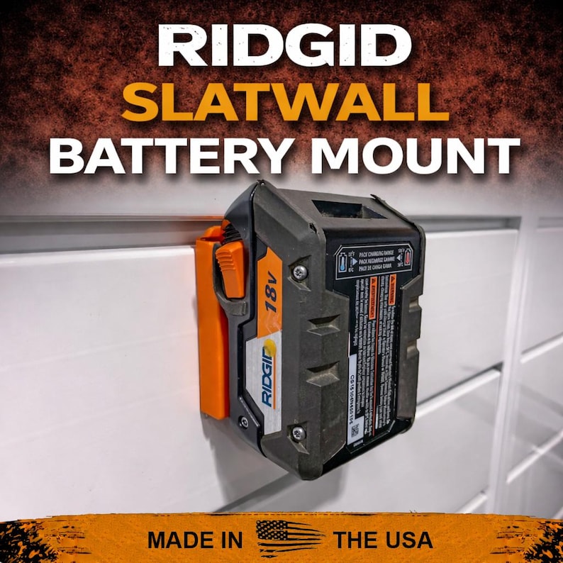 Puede incluir: Una bater&iacute;a Ridgid de 18V negra y naranja montada en una pared de lamas blanca. El soporte de la bater&iacute;a est&aacute; etiquetado como "RIDGID SLATWALL BATTERY MOUNT" y "MADE IN THE USA".