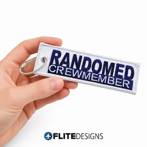 May include: A rectangular white embroidered fabric keychain featuring the dark blue text "RANDOMED CREWMEMBER" in a bold, sans-serif font. The tag includes a silver-toned metal eyelet and a flexible, circular wire cable fastener for attachment.