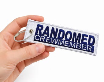 Puede incluir: Un llavero rectangular de tela bordada en blanco con el texto azul oscuro "RANDOMED CREWMEMBER" en una fuente sans-serif en negrita. La etiqueta incluye un ojal de metal plateado y un cierre de cable de alambre circular y flexible.