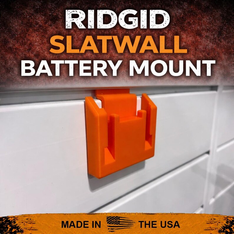 Puede incluir: Un soporte de bater&iacute;a Ridgid naranja para panel ranurado est&aacute; fijado a un panel ranurado blanco. El texto "RIDGID SLATWALL BATTERY MOUNT" est&aacute; en la parte superior de la imagen. La parte inferior de la imagen dice "MADE IN THE USA".