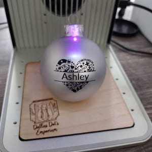 Könnte beinhalten: Ein silberner Weihnachtsbaumschmuck mit einem schwarzen Herzdesign und dem Namen "Ashley" eingraviert. Der Schmuck wird von innen beleuchtet und strahlt in einem violetten Licht. Der Schmuck liegt auf einem Holzsockel mit den eingravierten Worten "Dallas Dot's Emporium".